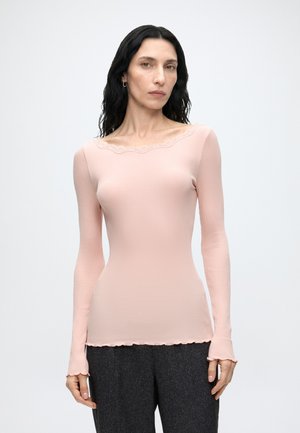 Frau mit langen schwarzen Haaren, die ein hellrosa eng anliegendes Langarmshirt und dunkelgraue Hosen trägt, steht vor einem schlichten hellen Hintergrund.