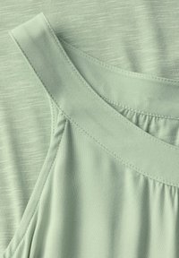 Světlá zelená látka s hladkým povrchem, má široké ramínka a detaily s nařasením. Jsou viditelné ploché švy a mírný lesk.