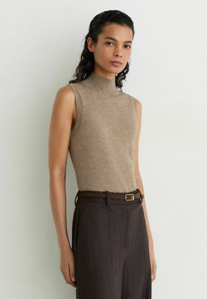 Jonge vrouw met donker krullend haar die een mouwloze beige coltrui draagt en een hoog uitgesneden donkerbruine broek met riem.