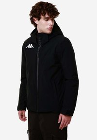 Giacca impermeabile nera con chiusura a zip, cappuccio regolabile e logo Kappa sul lato sinistro del petto. Presenta polsini testurizzati e una silhouette snella.
