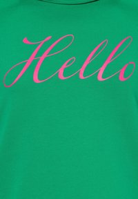 T-shirt in cotone verde con una grande stampa cursiva rosa "Hello" al centro. Tessuto morbido, design classico con collo tondo.