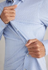 Jasnoniebieska koszula z guzikami, z geometrycznym wzorem, o gładkiej fakturze i białymi guzikami. Koszula jest częściowo rozpięta.