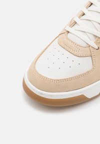 Baskets beige et blanches avec embout perforé, lacets blancs et semelle en gomme visibles sur fond blanc.