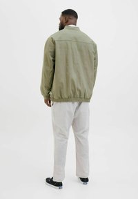 Olijfgroene bomberjack met ribbels aan de manchetten en onderrand, relaxed fit, lichtgewicht stof. Gecombineerd met lichtgrijze broek en zwarte schoenen.
