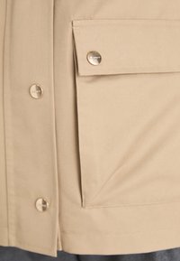 Beige jacket featuring a button closure and a front pocket with a metallic snap, made of smooth fabric with a slight sheen.