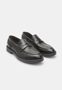 Mocasines de cuero negro con una correa decorativa, detalles cosidos y un tacón bajo cuadrado. Textura suave con un diseño de punta redondeada.