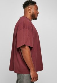 Burgundská oversized tričko, bavlněný materiál, krátké rukávy, kulatý výstřih, volný střih, boční pohled, spárované se šedými džínami.