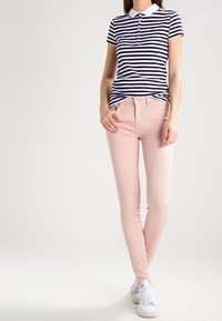 Camisa polo a rayas en azul marino y blanco con cuello blanco, combinada con jeans skinny en rosa claro y zapatillas blancas. Diseño sencillo, silueta ajustada.
