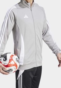 Graue zip-off Sportjacke mit weißen Akzenten und drei vertikalen Streifen, kombiniert mit schwarzen Hosen; hält einen mehrfarbigen Fußball.