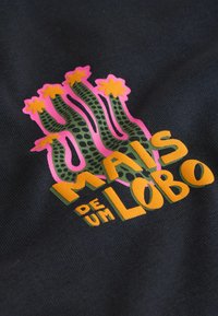 Grüner gesprenkelter Kaktus mit rosa Umriss und gelben Blüten über fettem gelbem Text "Mais de um Lobo" auf schwarzem Stoffhintergrund.