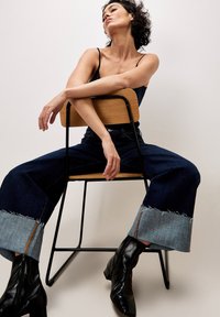 Weite Denim-Jeans mit dunkler Waschung und hellem Denim-Cuff, kombiniert mit schwarzen Ankle-Boots. Modell sitzt auf einem Holzstuhl.