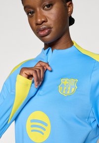 Μπλε φανέλα μακρυμάνικη με κίτρινα στοιχεία, με φερμουάρ και κεντημένο λογότυπο συλλόγου στο στήθος, καθώς και κίτρινο λογότυπο Spotify.