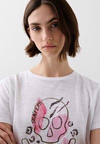 White cotton t-shirt featuring a graphic print of a skull with pink accents and black details. Short sleeves and a relaxed fit.