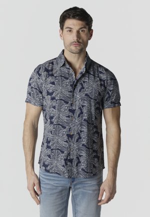 Hombre vistiendo una camisa azul marino de manga corta con botones y patrones de hojas blancas, y jeans azul claro, de pie contra un fondo liso.