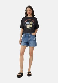 T-shirt noir surdimensionné avec imprimé graphique, associé à un short en denim bleu déchiré. La mannequin porte des sandales à plateforme noires.