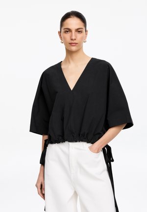 Kobieta z ciemnymi włosami zaczesanymi do tyłu, ubrana w czarną bluzkę z dekoltem w serek i szerokimi rękawami oraz białe spodnie z wysokim stanem, stoi z jedną ręką w kieszeni.