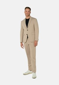Beige Zweiteiler-Anzug mit einreihiger Blazerjacke und geraden Hosen, kombiniert mit einem schwarzen Hemd und weißen Sneakern. Glatte Textur.
