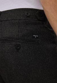 Pantaloni in denim nero con una finitura leggermente texture, dotati di chiusura a bottone, tasca posteriore e una piccola etichetta del marchio.