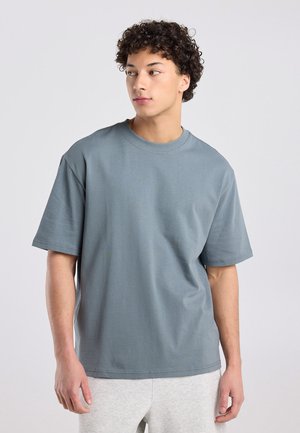 Jonge man met krullend haar, gekleed in een loszittend grijs T-shirt en lichtgrijze broek, kijkt naar links tegen een effen achtergrond.