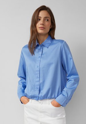 Vrouw met bruin haar, gekleed in een blauw overhemd met knopen en witte broek, staand met handen in de zakken tegen een grijze achtergrond.