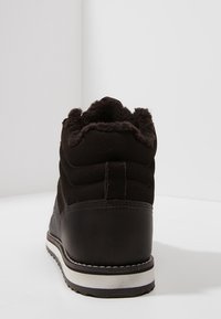 Bota de invierno negra con forro de piel, parte superior de tejido acolchado y suela con ribete blanco, vista desde atrás sobre una superficie blanca.