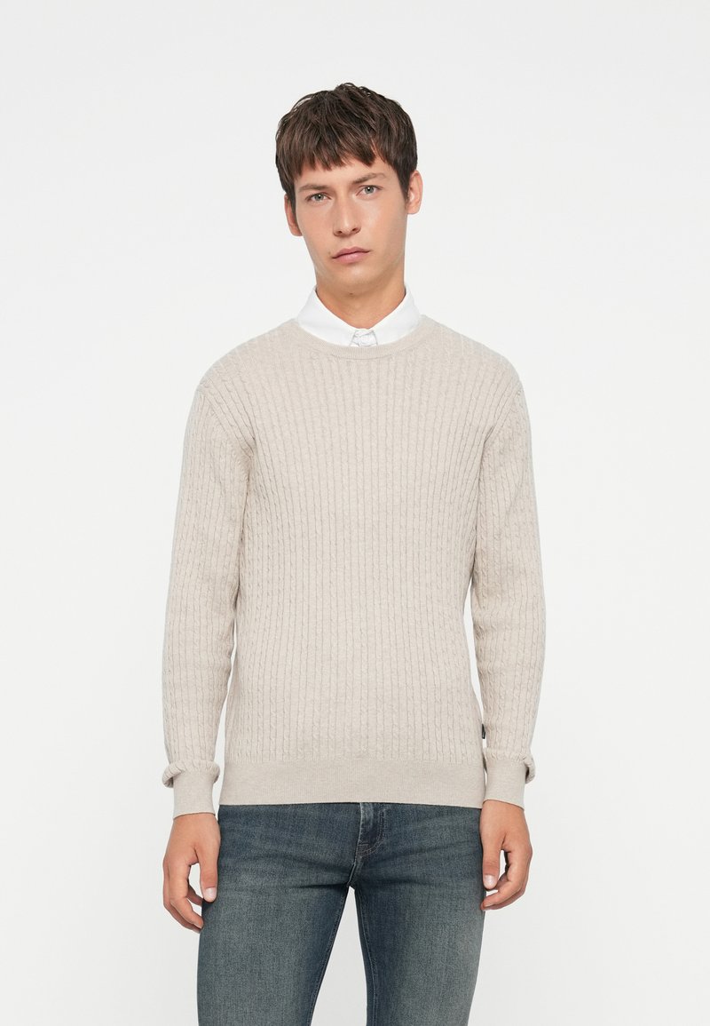 Lys beige genser med ribbet tekstur, rund halsutringning og lange ermer, sammen med mørk denimjeans. Enkel og uformell design.