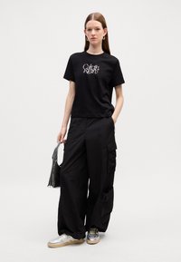 T-shirt en coton noir avec un texte graphique blanc, associé à un pantalon cargo noir ample avec des poches, et des baskets argentées avec une semelle beige.