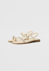 Sandales plates beiges avec des brides entrecroisées et une fine bande ornée. La semelle est plate et faite d'un matériau doux et texturé.