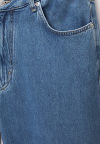 Tissu de jean en denim bleu avec poche avant visible, passants de ceinture et détails de bouton en laiton en vue rapprochée.