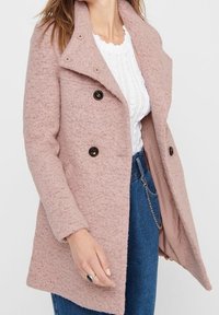Cappotto in lana rosa, testurizzato, con rever a scollatura, bottoni doppi e tasche laterali, indossato sopra un top in maglia bianca e jeans.