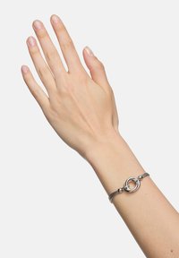 Srebrna bransoletka z przeplatającymi się okrągłymi ogniwami i gładką metalową bandą, noszona na dłoni z naturalnymi paznokciami na jednolitym tle.