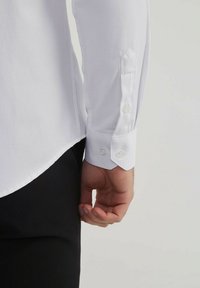 Chemise blanche en tissu texturé, avec un col pointu, des poignets à double bouton et un ourlet arrondi, associée à un pantalon foncé.