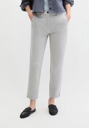 Osoba ubrana w jasnoszare wąskie spodnie, czarne wsuwane mokasyny i ciemnoszarą kurtkę, z rękami w kieszeniach, stojąca na białym tle.