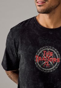 Zwarte katoenen T-shirt met een ronde halslijn, voorzien van een rode grafische print met de tekst "DE TRAITORS" binnen een circulair ontwerp. Textuurstof.