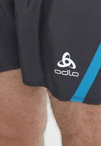 Pantalones cortos deportivos negros con una franja de acento turquesa, que presentan el logo blanco de Odlo en la parte inferior izquierda y una tela suave y texturada.