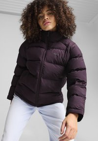 Lilla puffer-jakke med høj krave, lynlås lukning og syede paneler. Har et sort logo på brystet og er parret med lyse jeans.
