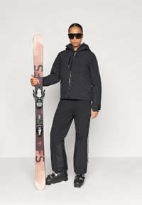 Schwarze Skijacke mit Kapuze; passende schwarze Hose mit weißen seitlichen Streifen; schwarze Skischuhe; hält pinke und braune Ski.