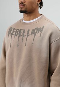 Beige sweatshirt met een ronde halslijn en doorgezakte schouders. Heeft een fonkelende zilveren "REBELLOON"-grafiek met een druppel-effect aan de voorkant.