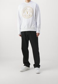 Vit sweatshirt med guldbroderad logotyp. Parat med svarta byxor och vita sneakers. Avslappnad passform med ribbade muddar och fåll.