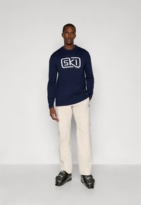 Homem vestindo suéter azul-marinho com o logotipo "SKI", calças bege e botas de esqui pretas, em pé contra um fundo cinza claro simples.