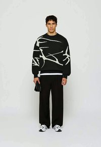 Junger Mann, der einen Pullover mit schwarz-weißen abstrakten Mustern, schwarze weit geschnittene Hosen, weiße Sneakers trägt und schwarze Handschuhe hält, vor einem schlichten Hintergrund steht.
