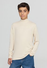 Beżowa koszula z długim rękawem i wysokim kołnierzem, wykonana z miękkiego materiału. Posiada luźny krój i subtelne marszczenia. Połączona z niebieskimi szortami dżinsowymi.