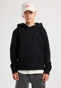 Felpa nera con cappuccio, dotata di tasca a marsupio e un piccolo logo sul petto. Indossata sopra una maglietta bianca e abbinata a pantaloni grigi. Cappello beige.