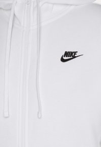 Bílá mikina na zip vyrobená z měkkého materiálu, která má černě vyšívané logo Nike na levé straně hrudi a nastavitelnou kapuci se šňůrkou.