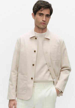 Mand iført beige jakke med knapper over en lys sweater med hånden i lommen, stående mod en ensfarvet hvid baggrund.