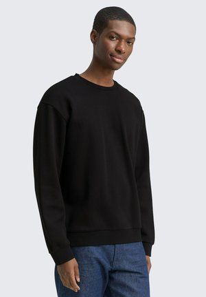 Junger Mann trägt einen schlichten schwarzen Sweatshirt mit Rundhalsausschnitt und langen Ärmeln und blaue Jeans, steht vor einem hellgrauen Hintergrund.