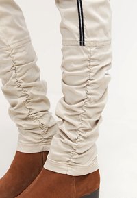 Beżowe botki damskie z sztucznej skóry, sięgające kostki, z marszczonym wzorem i czarnymi paskami po bokach. Brązowy zamszowy obcas o gładkiej teksturze.