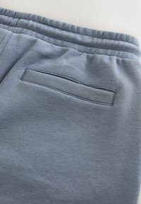 Gros plan sur un pantalon de survêtement gris à taille élastique montrant une poche arrière fendue horizontale avec une couture renforcée.