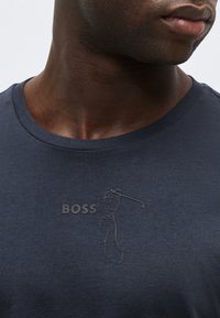 Άνδρας με σκουρόχρωμο δέρμα που φοράει μπλε-σκούρο πουκάμισο πόλο με υφή και τη λέξη "BOSS" εγχάρακτη στο στήθος.