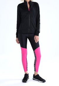 Černá zipová bunda s vysokým límcem, spárovaná s černými a růžovými legínami se geometrickým vzorem, označená logem značky po straně.